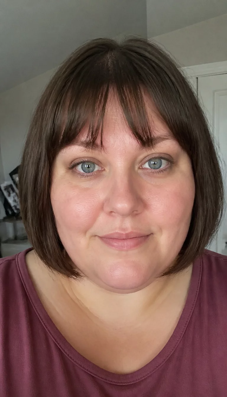Polished Brunette Bob With Blunted Fringe casual photo of a 31-year-old plus size Caucasian woman with a chubby face and a straight, petite nose. Her face skin has smooth, visible pores. The eyes are blue. She is wearing a simple top with a chin-length brown bob and bangs hairstyle, front view