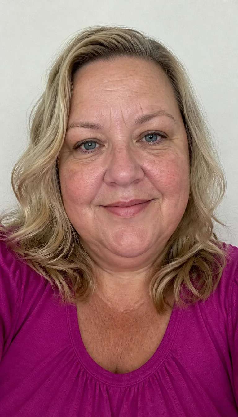The Soft Blonde Wave selfie of a 44-year-old plus size Caucasian woman with a chubby face and a rounded, soft nose. The skin is heavily textured with a complex pattern of light sunspots on the cheeks and a faint, thin scar traversing the left eyebrow. The eyes are deep-set blue. She is wearing a magenta top with a blonde, wavy, shoulder-length hairstyle, front view, plain white wall.