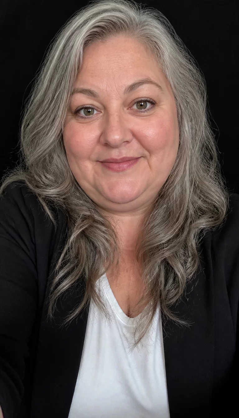 selfie of a 52-year-old plus size Caucasian woman with a chubby face and a button nose. The eyes are wide set hazel. She is wearing a black blazer over a white t-shirt with long, wavy, grey hair, front view, dark background.