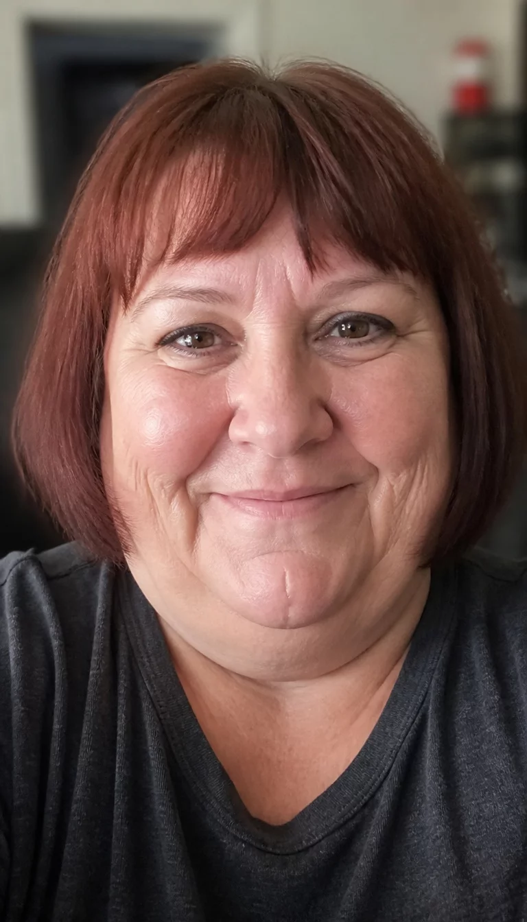 The Crimson Bob With Fringe selfie of a 55-year-old plus size Caucasian woman with a chubby face and a delicate, narrow nose. The skin is heavily textured with a complex pattern of deep laugh lines and a faint scar on the chin. The eyes are deep set brown. She is wearing a dark grey t-shirt with a short, reddish-brown bob with bangs, front view, blurred background.