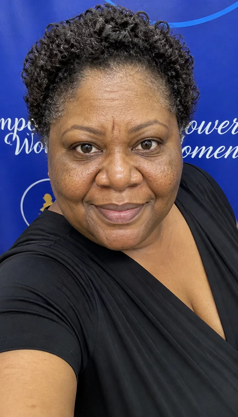 Classic Dark Curls selfie of a 53-year-old plus size Black woman with a chubby face and a slightly flared nose. The skin is heavily textured with a complex pattern of uneven skin tone and a small permanent crease on the forehead. The eyes are large, dark brown. She is wearing a black dress with short, dark curly hair, front view, blue banner background.