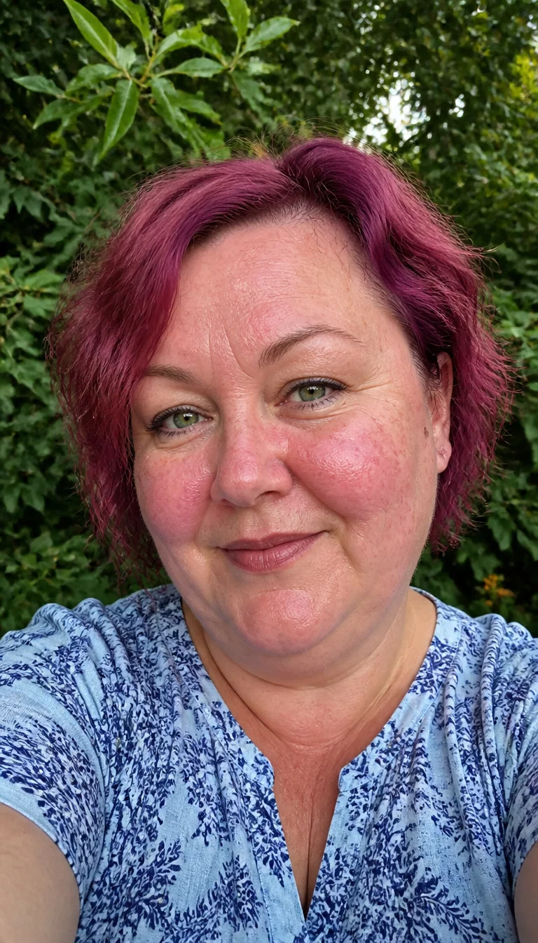 The Playful Pink Wave selfie of a 51-year-old plus size Caucasian woman with a chubby face and a nose with a small bridge bump. The skin is heavily textured with a complex pattern of visible capillaries on the cheeks and a small birthmark near the ear. The eyes are bright green. She is wearing a light blue patterned top with a short, wavy, dark pink bob, front view, background of green foliage.