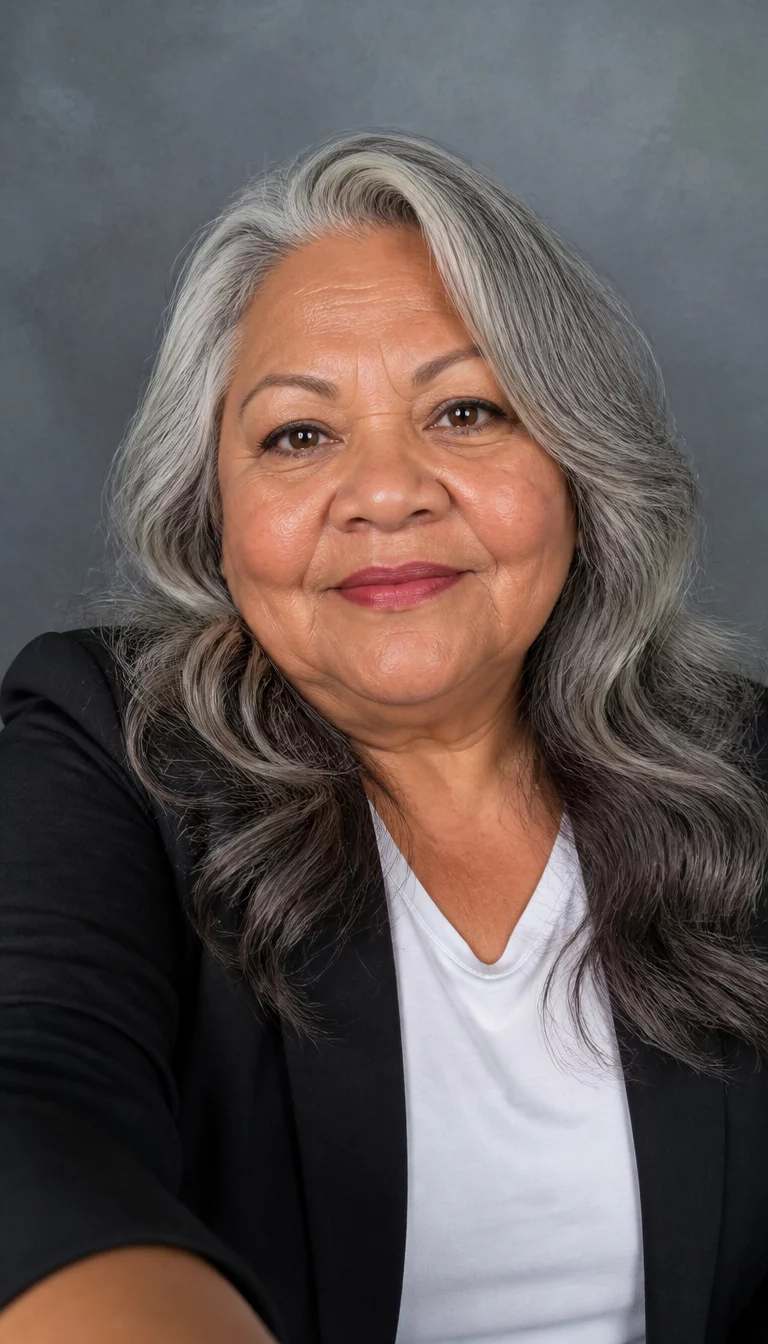 Striking Silver Waves selfie of a 67-year-old plus size Latina woman with a chubby face and wider nose bridge. Her face skin has sun-kissed texture. The eyes are warm brown. She is wearing a black blazer over a white t-shirt with a long grey hair styled in loose waves hairstyle, front view, professional studio background.