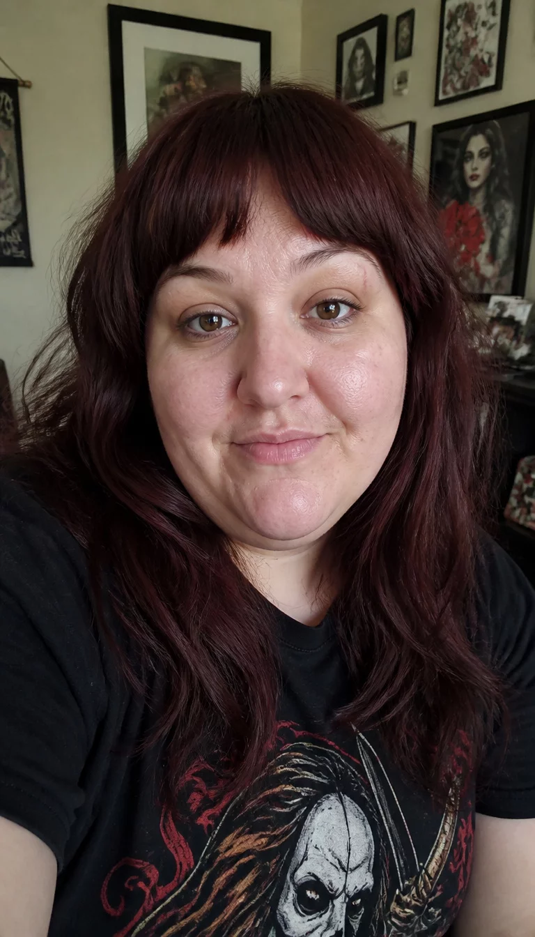 selfie of a 32-year-old plus size Caucasian woman with a chubby face and a slightly crooked nose. The skin is heavily textured with a complex pattern of pale skin and a faint scar above the right eye. The eyes are dark brown. She is wearing a black t-shirt with a graphic with a long, dark reddish-brown hair styled in layers with bangs hairstyle, front view, room with framed pictures.