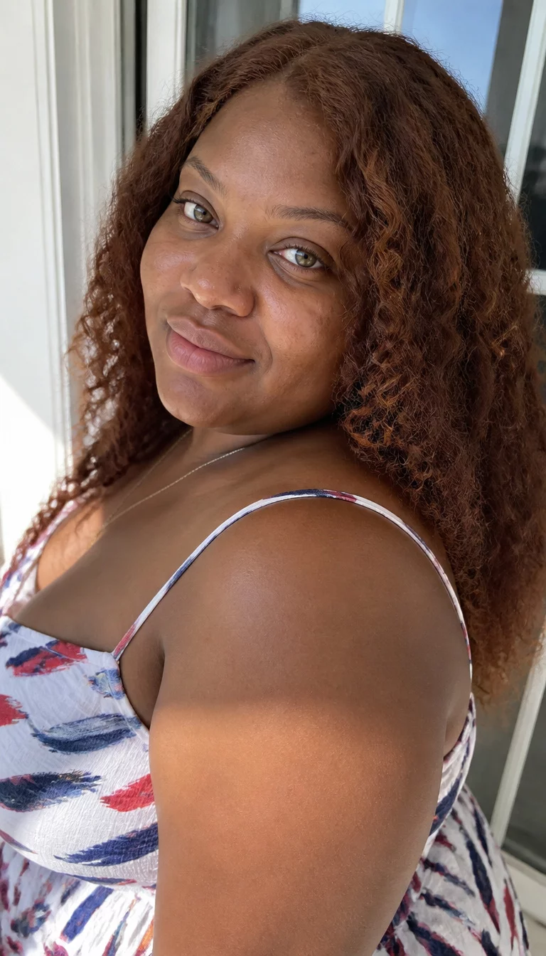 casual photo of a 29-year-old plus size Black woman with a chubby face and a rounded nose. The skin is heavily textured with a complex pattern of faint acne scarring on the chin and a few dark spots on the forehead. The eyes are large and honey colored. She is wearing a white spaghetti strap dress with a blue and red abstract design with voluminous, reddish-brown curly hair that falls past the shoulders, three-quarter side view, bright natural light.
