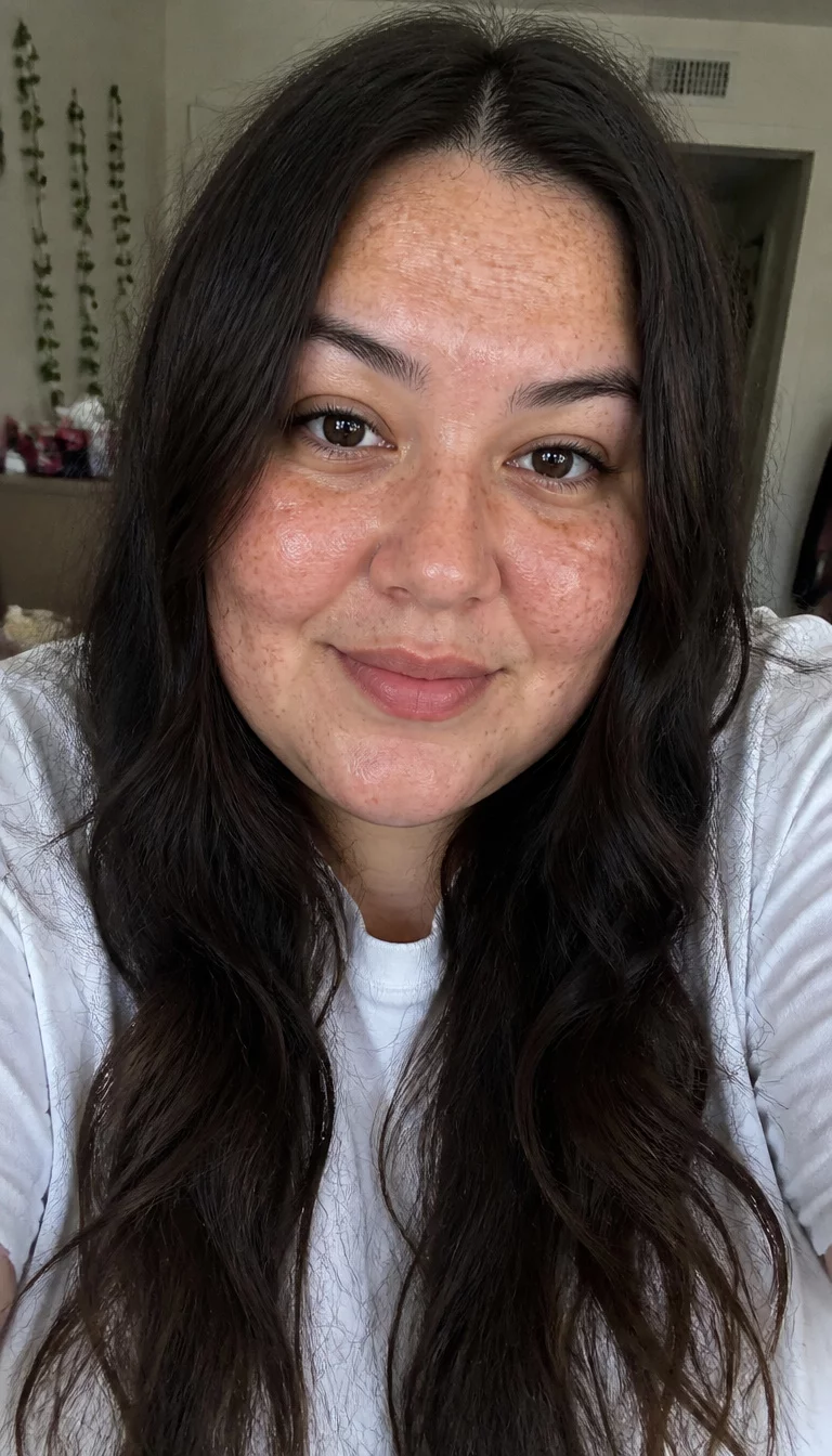selfie of a 29-year-old plus size woman with a chubby face and a straight nose. The skin is heavily textured with a complex pattern of freckles and a small blemish on the forehead. The eyes are dark brown. She is wearing a white t-shirt with long, dark, wavy brown hair, front view, indoor background.