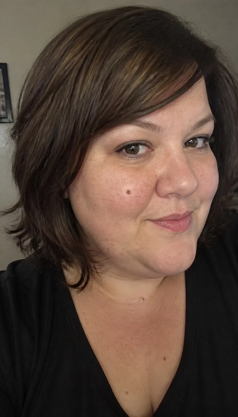 Side Swept Brunette Bob selfie of a 35-year-old plus size Caucasian woman with a chubby face and a rounded nose. The skin is heavily textured with a complex pattern of small moles and a faint, thin scar traversing the left eyebrow. The eyes are deep-set brown. She is wearing a dark top with a dark brown layered bob hairstyle with side-swept bangs, three-quarter view, soft indoor lighting.