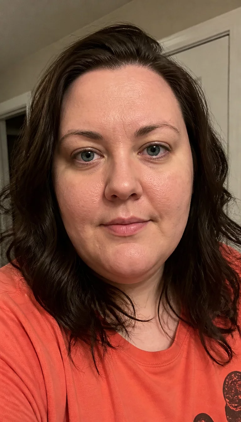 Vibrant Waves in Autumn Orange selfie of a 29-year-old plus size Caucasian woman with a chubby face and a straight nose. The skin is heavily textured with a complex pattern of light acne scarring and a faint, thin scar traversing the left eyebrow. The eyes are deep-set blue. She is wearing an orange shirt with a dark brown wavy shoulder length hairstyle, front view, close-up indoor background.