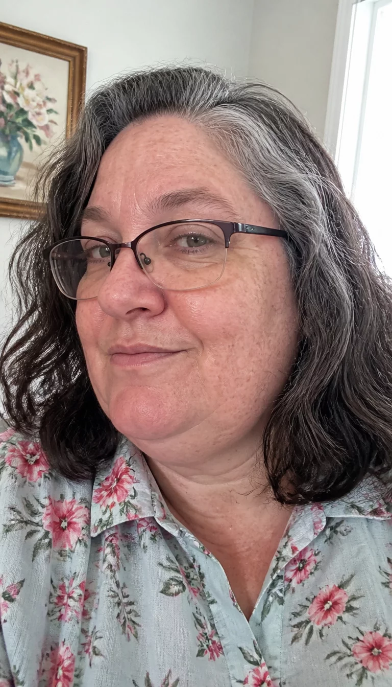Salt and Pepper Sophistication selfie of a 47-year-old plus size Caucasian woman with a chubby face and a wide nose. The skin is heavily textured with a complex pattern of age spots and a faint, thin scar traversing the left eyebrow. The eyes are deep-set gray. She is wearing a floral collared shirt with a gray and black wavy medium hairstyle with natural texture and glasses, side view, brightly lit room.
