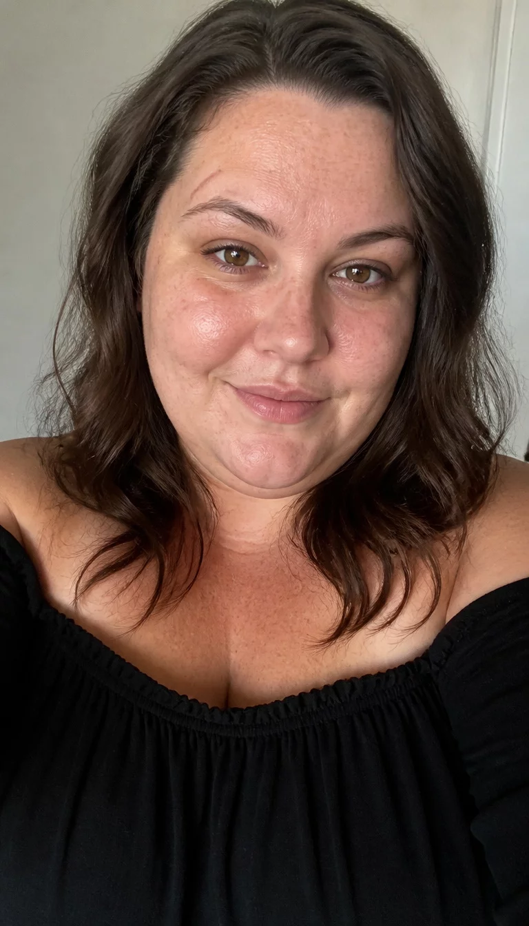Off The Shoulder Brunette Waves selfie of a 33-year-old plus size Caucasian woman with a chubby face and a straight nose. The skin is heavily textured with a complex pattern of light freckles and a faint, thin scar traversing the left eyebrow. The eyes are deep-set brown. She is wearing a black off-the-shoulder top with a dark brown wavy shoulder length hairstyle, front view, simple indoor background.
