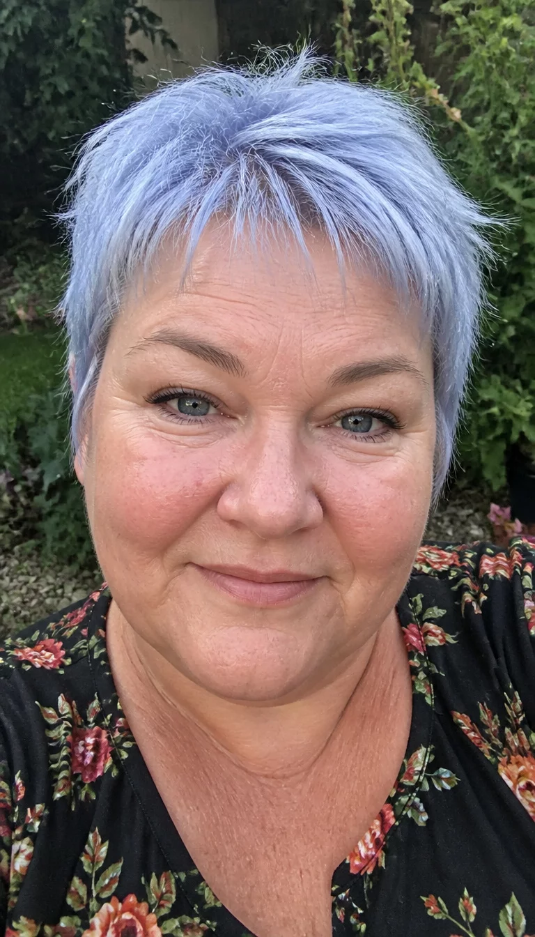 selfie of a 44-year-old plus size Caucasian woman with a chubby face and a broad, soft nose. The skin is heavily textured with a complex pattern of fine lines and a small patch of redness near the nose. The eyes are blue-gray. She is wearing a dark floral top with a short choppy pixie cut in a light silver-blue shade, front view, natural garden background.