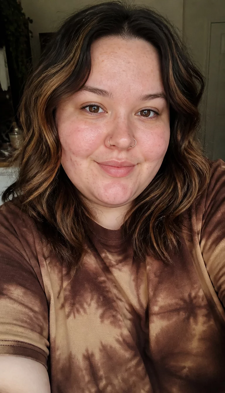 Caramel Highlighted Waves selfie of a 28-year-old plus size fair complexion woman with a chubby face and a nose ring on a rounded nose. The skin is heavily textured with a complex pattern of light acne and some freckles. The eyes are brown. She is wearing a brown tie-dye t-shirt with medium-length wavy hair in a dark base with caramel-colored highlights hairstyle, front view.