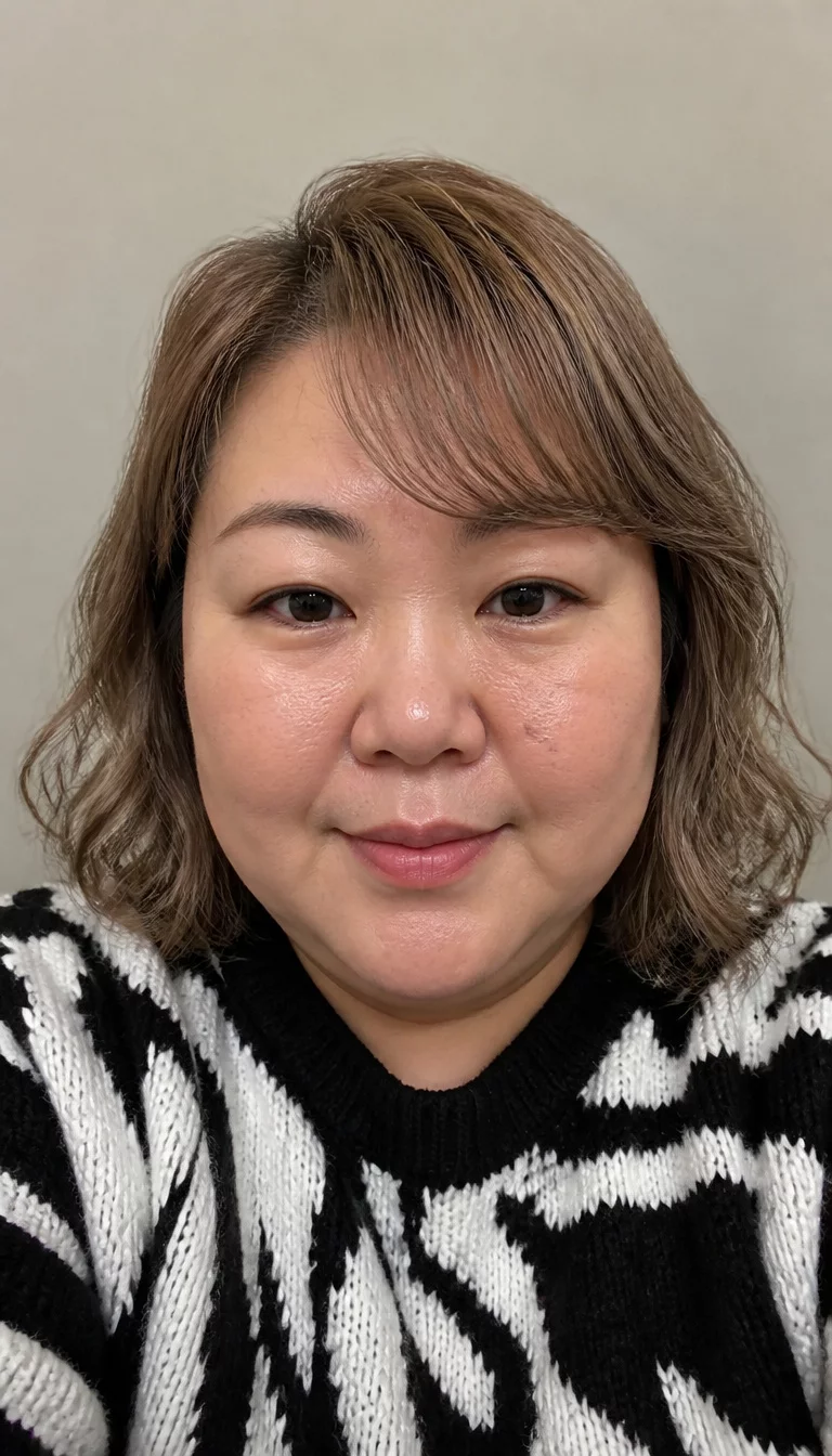 Soft Waves and Side Swept Bangs selfie of a 42-year-old plus size East Asian woman with a chubby face and a small, button nose. The skin is heavily textured with a complex pattern of enlarged pores on the nose and a faint beauty mark on the cheek. The eyes are dark brown. She is wearing a black and white patterned sweater with a short, dark blonde wavy hair with side part and bangs hairstyle, front view, neutral background.