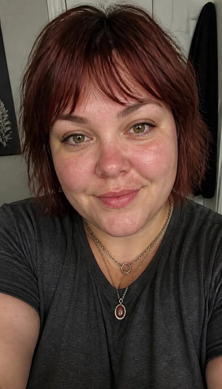 Choppy Mahogany Bob with Necklaces selfie of a 30-year-old plus size fair complexion woman with a chubby face and a button nose. The skin is heavily textured with a complex pattern of redness on the nose and a few freckles. The eyes are hazel. She is wearing a dark gray t-shirt and layered necklaces with a reddish-brown, choppy bob with textured bangs hairstyle, front view.